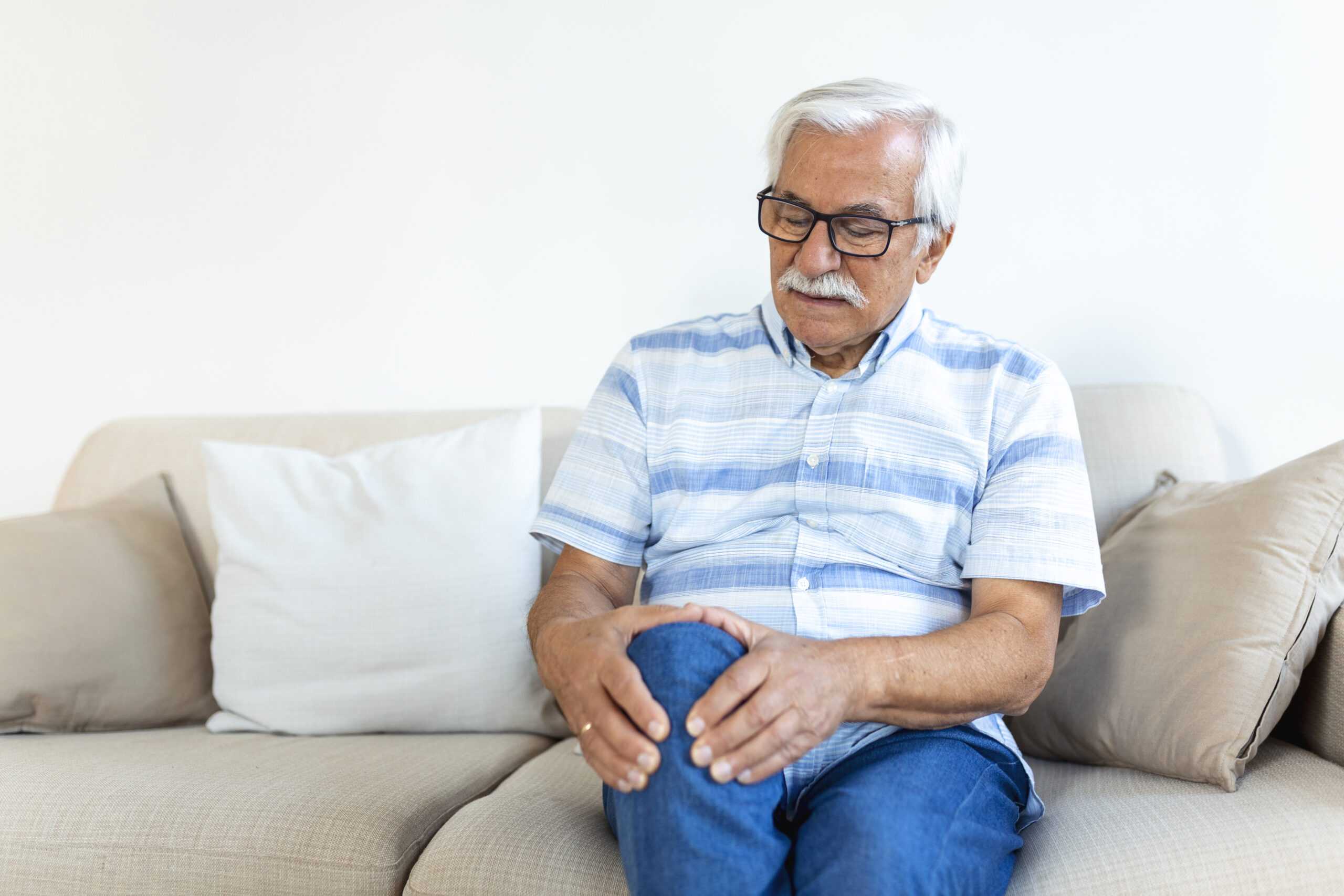 elderly man sitting sofa home touching his painful knee people health care problem concept unhappy senior man suffering from knee ache home scaled
