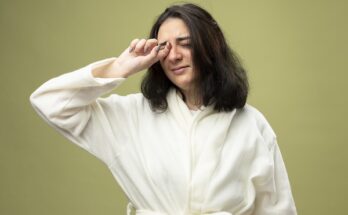 weak young caucasian ill girl wearing robe wiping eye with closed eyes isolated olive green background